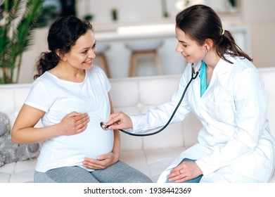 Health of the pregnant woman and baby. Female friendly doctor examines pregnant mixed race smiling woman, listening to baby's heartbeat, sitting on a sofa at home or hospital,healthy pregnancy concept