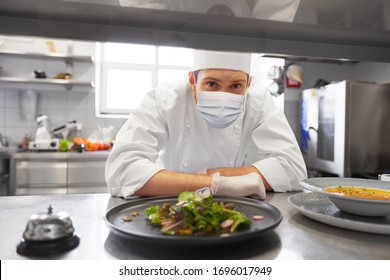 health, safety and pandemic concept - male chef cook wearing face protective medical mask for protection from virus disease with plate of soup and salad at restaurant kitchen
