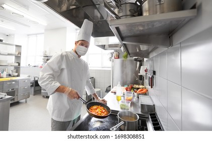 health, safety and pandemic concept - male chef cook wearing face protective medical mask for protection from virus disease cooking food in frying pan at restaurant kitchen - Powered by Shutterstock