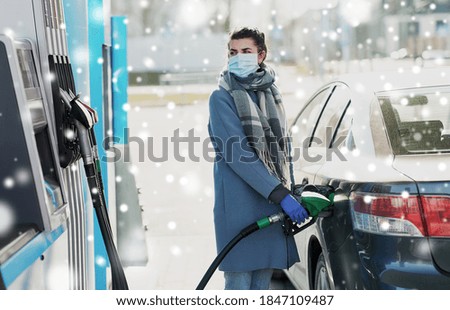 health, safety and pandemic concept - young woman wearing protective medical mask and gloves filling her car with gasoline at gas station over snow