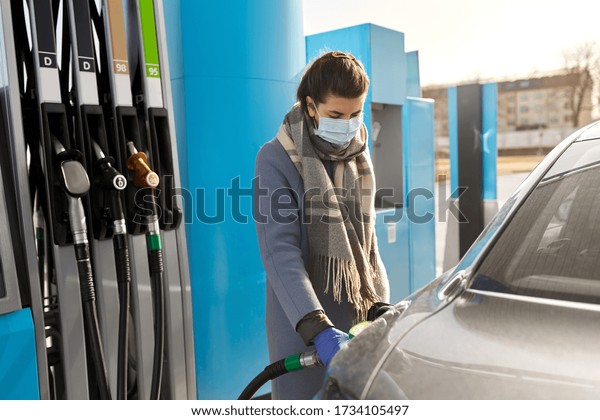 health, safety and pandemic concept - young woman\
wearing protective medical mask and gloves filling her car with\
gasoline at gas station
