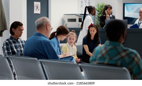 Health Specialist Talking To Small Child And Mother In Hospital Reception Lobby, Taking Notes About Consultation. Nurse Consulting Little Girl Sitting In Facility Waiting Room, Healthcare Support.