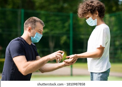 Health And Sport Concept. Father In Face Mask Using Sanitizer With His Son, Spraying On Child's Hands In The Playground