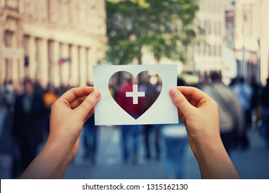 Health And Wellbeing Global Issue As Human Hands Holding A Paper Sheet With Heart And Cross Icon Over A Crowded Street Background. Healthcare Medical Insurance, Good Life Concept.