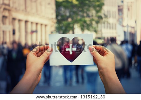 Health and wellbeing global issue as human hands holding a paper sheet with heart and cross icon over a crowded street background. Healthcare medical insurance, good life concept.