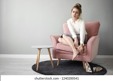 Health, Wellbeing, Injury And Lifestyle Concept. Indoor Shot Of Blonde Young Woman Doing Foot Massage Because Of Working On Her Feet All Day. Sore Foot, Sprained Ankle, Heel And Toe Callus