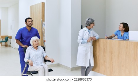 Healthcare assistant pushes wheelchair with senior woman in motion along corridor of hospital near reception desk with on-call nurse and doctor. Patient care in hospital - Powered by Shutterstock