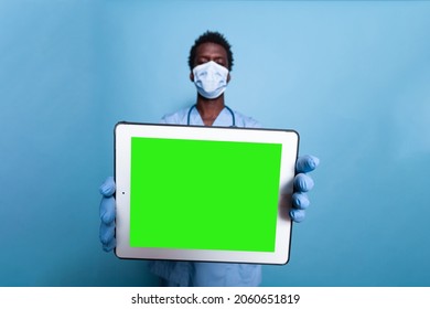 Healthcare Assistant Showing Tablet With Horizontal Green Screen. Medical Nurse With Face Mask, Gloves And Stethoscope Holding Gadget With Chroma Key And Mockup Template For Isolated Background.