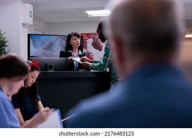 Healthcare Center Receptionist Holding Clipboard For Man To Sign Before Doctor Appointment In Busy Waiting Room. African American Patient Signing Admission Form Into Clinic At Front Desk Reception.