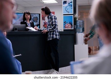 Healthcare Center Receptionist Holding Clipboard For Asian Patient To Sign Before Doctor Appointment In Busy Waiting Room. Young Man Signing Admission Form Into Clinic At Front Desk Reception.