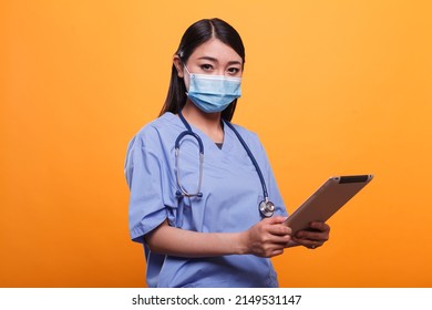 Healthcare Clinic Asian Nurse Wearing Virus Protection Facemask And Stethoscope While Having Modern Tablet. Caretaker Woman Holding Touchscreen Pad While Wearing Protective Mask On Orange Background.