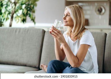 Healthcare concept. Mature caucasian blonde dressed in a casual wear sits on the couch in a living room, holding a glass of pure water in a hand and smiling, follow healthy lifestyle