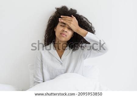 Healthcare concept. Sick afro american woman with closed eyes touch forehead, suffering from headache, sitting on bed in pajamas. Young female feeling unhealthy, strong sudden pain, morning hangover
