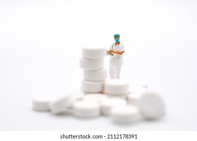 Healthcare And Medical Concept. Closeup Of Woman Doctor Miniature Figure With Patient File Looking To Stack Of White Pills On White Background.