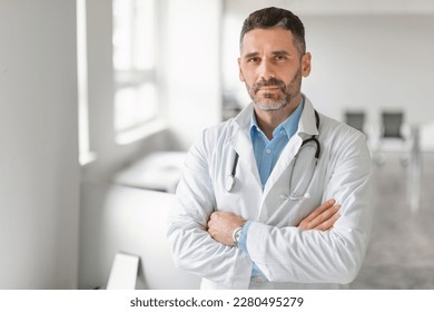 Healthcare and medicine concept. Portrait of confident middle aged male doctor with folded arms posing and looking at camera, standing in clinic office interior, copy space - Powered by Shutterstock