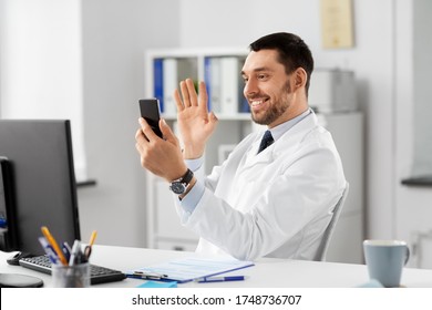 Healthcare, Medicine And People Concept - Smiling Male Doctor With Smartphone Having Video Call And Waving Hand At Hospital