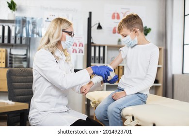 Healthcare And Orthopedics Concept. Mature Professional Blond Female Doctor Imposing Bandage On Teen Boy's Hand, Suffering From Pain, In Modern Medical Cabinet