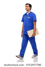 Healthcare, Profession And Medicine Concept - Happy Smiling Indian Doctor Or Male Nurse In Blue Uniform With Clipboard And Stethoscope Walking Over White Background