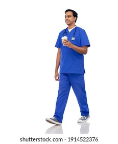 Healthcare, Profession And Medicine Concept - Happy Smiling Indian Doctor Or Male Nurse In Blue Uniform With Takeaway Coffee Cup And Stethoscope Walking Over White Background