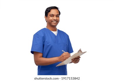 Healthcare, Profession And Medicine Concept - Happy Smiling Indian Doctor Or Male Nurse In Blue Uniform Writing Medical Report On Clipboard Over White Background