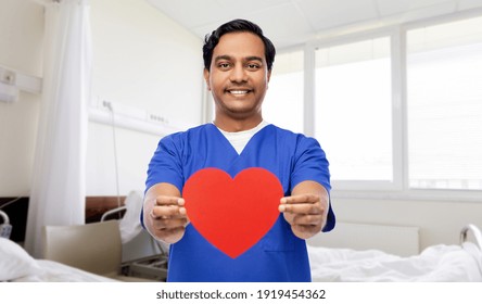 Healthcare, Profession And Medicine Concept - Happy Smiling Indian Doctor Or Male Nurse In Blue Uniform With Red Heart Over Corridor At Hospital On Background