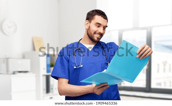 healthcare, profession\
and medicine concept - happy smiling doctor or male nurse in blue\
uniform reading medical report in folder over medical office at\
hospital on\
background