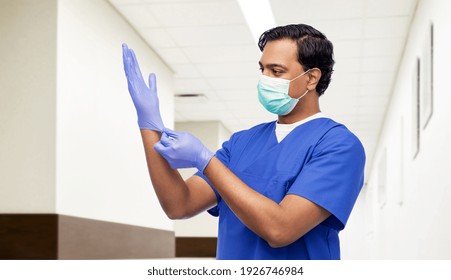 healthcare, profession and medicine concept - indian doctor or male nurse in blue uniform and face protective medical mask putting surgical gloves on over hospital corridor background