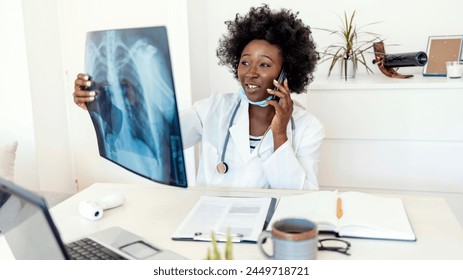 Healthcare, technology, people and medicine concept - young female doctor in white coat with laptop computer looking at x-ray in medical office while talking via phone. Female doctor reviews x-ray. - Powered by Shutterstock