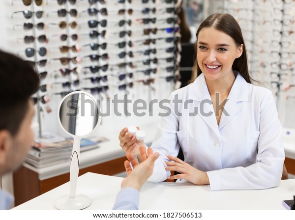 Healthcare And\
Vision Correction Concept. Happy female doctor optometrist giving\
plastic container with contacts lens to man customer sitting at\
desk. Consultation at the optics\
store