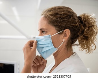 A healthcare worker adjusts a light blue mask while in a clinical environment, demonstrating proper face covering practices for safety and health. - Powered by Shutterstock