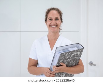 A healthcare worker with a big smile stands confidently in a clinic, holding a folder filled with patient records. She is dressed in professional attire, ready for work. - Powered by Shutterstock