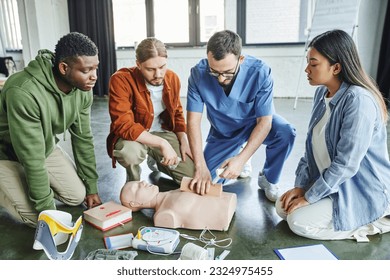 healthcare worker tamponing wound on simulator with bandage while showing life-saving skills to multicultural team near CPR manikin, defibrillator and medical equipment, emergency response concept