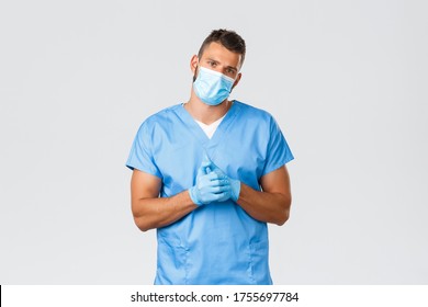 Healthcare Workers, Covid-19, Coronavirus And Preventing Virus Concept. Handsome Tired Doctor, Male Nurse In Medical Mask And Scrubs, Crack Knuckles And Look Camera Exhausted, Grey Background