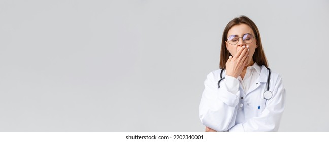 Healthcare workers, medicine, insurance and covid-19 pandemic concept. Young doctor in white scrubs, female physician working hard night shift during coronavirus, yawning and cover mouth - Shutterstock ID 2202340001