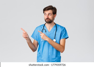 Healthcare Workers, Pandemic And Coronavirus Outbreak Concept. Uncertain And Indecisive Bearded Doctor, Physician Or Nurse In Scrubs, Stethoscope, Smirk, Looking Pointing Left Curious