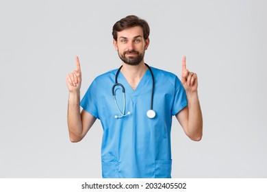 Healthcare workers, pandemic and coronavirus outbreak concept. Handsome bearded doctor in scrubs, stethoscope, pointing fingers up, showing top advertisement, treat patients from covid-19