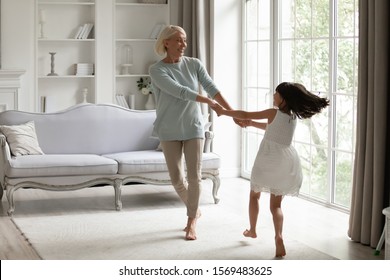 Healthy Active 50s Grandmother Holding Hands Little Granddaughter Dancing Or Spinning Standing Barefoot In Modern Warm Living Room Enjoy Priceless Time Together At Funny Weekend Activities Concept