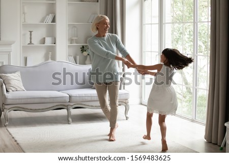 Healthy active 50s grandmother holding hands little granddaughter dancing or spinning standing barefoot in modern warm living room enjoy priceless time together at funny weekend activities concept