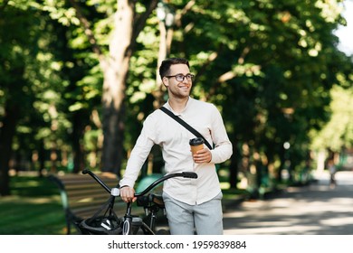 Healthy And Active Lifestyle, Going To Work On Eco Transport. Smiling Attractive Millennial Male Employee In Glasses With Bag And Cup Of Takeaway Coffee, Moving Bike In City Park In Summer, Outdoor