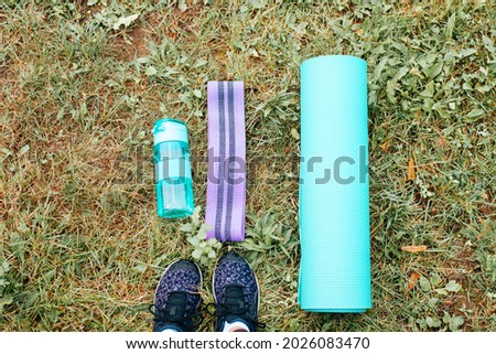 Healthy active lifestyle, sport concept. Yoga mat, elastic expander, bottle of water and female legs in sneakers on green grass top view, outdoors. First-person view of outdoor training equipment.