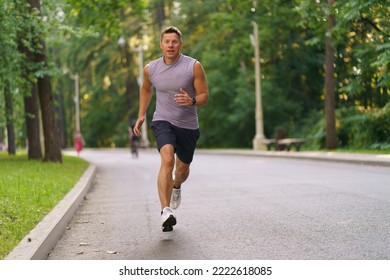 Healthy active man runner running in park. Handsome young male jogger trains cardio going for a run in city park. Lush green foliage of trees, road in the background in summer. He is looks in camera - Powered by Shutterstock
