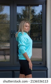 Healthy Activity. Blonde Girl In Blue Hoodie And Black Cycling Shorts Is Standing In Profile Near Park Cafe Background And Looking Away With Smile. Sport Lifestyle Concept, Free Space