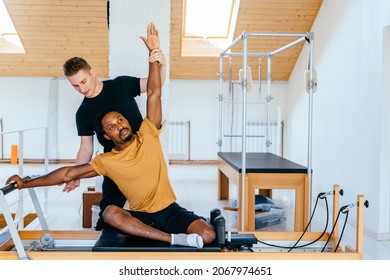 Healthy african man practicing pilates in exercise studio on reformer special equipment. Male trainer therapist helping patient doing exercises on reformer pilates machine. Balance control concept.
