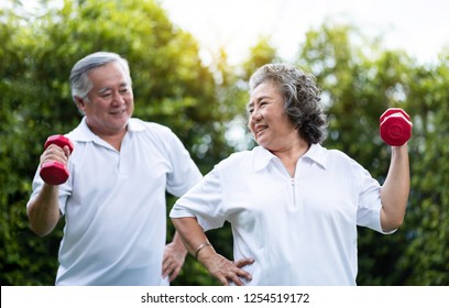 Healthy Asian Elderly Couple Exercising Training With Red Dumbbells Over Green Nature Togetherness. Happy Smiling Chinese Old Man And Woman Relaxing During Workout In The Morning. Strength, Wellness 