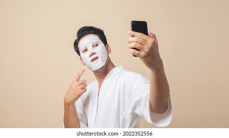 Healthy Asian Man In Bathrobe With Facial Sheet Face Mask For Skincare He Use Smartphone Take A Selfie On Brown Color Background.