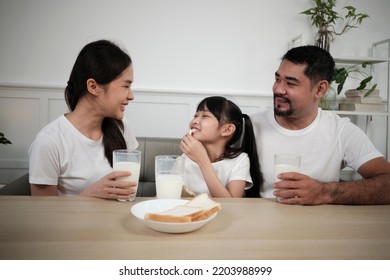 A Healthy Asian Thai Family, A Little Daughter, And Young Parents Drink Fresh White Milk In Glass And Bread Joy Together At A Dining Table In Morning, Wellness Nutrition Home Breakfast Meal Lifestyle.