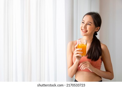 Healthy asian woman drinking a glass of orange juice for refreshment while wearing sportswear for nutrition and vitamin c supplements at the window 