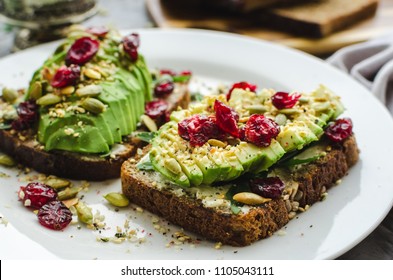 Healthy avocado toasts for breakfast or lunch with rye bread, cream cheese, arugula, sliced avocado, dried cranberry, pumpkin, hemp and sesame seeds. Vegetarian sandwiches. Clean eating concept.
