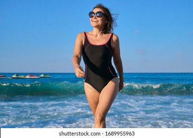 Healthy Beautiful Middle Aged Woman In Sunglasses Swimsuit Walking Along Seashore. Smiling Happy Female Relaxing In Beach Resort, Blue Sky Waves Sunset Background