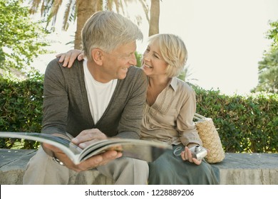 Healthy Beautiful Senior Tourist Couple Relaxing In Sunny Park Reading Travel Guide Book, Smiling Outdoors. Mature Man And Woman On Vacation Trip, Active Leisure Recreation Lifestyle.